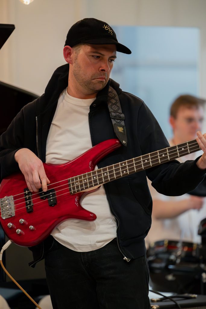 Man in a black cap and jacket playing a red bass guitar, focused on the fretboard during a rehearsal.
