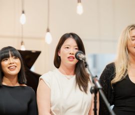 Three women singing on stage with microphones, smiling and performing together.