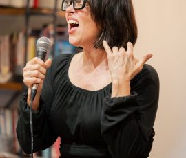 Woman wearing black dress sings into a microphone during a performance, glasses framing her face.