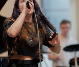 Female singer with purple-tinted hair sings into a handheld microphone on stage, blurred musician in the background