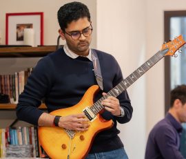 Man in glasses playing an orange electric guitar in a library or bookstore setting with shelves in the background.