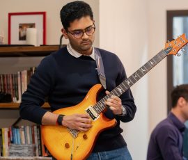 Man with glasses playing an orange electric guitar in a room with bookshelves in the background.