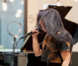 Female singer with purple-toned hair sings into a microphone in a studio with a grand piano in the background.