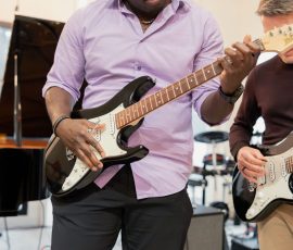 Man in a lavender shirt plays a black and white electric guitar, focused on the fretboard in a rehearsal space, with another guitarist nearby.