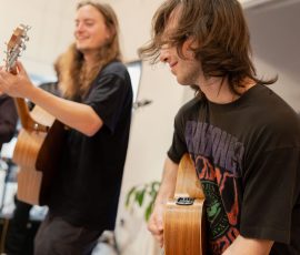 Two guitarists playing acoustics indoors, foreground musician with long hair wearing a black shirt and orange-brown pants, smiling as he strums.