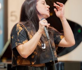 Female singer performing, holding a microphone with eyes closed, a piano visible in the background.