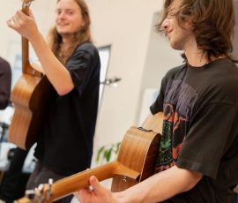 Two guitarists playing acoustic guitars in a bright indoor space; one foreground player in a black shirt smiling at the camera, the other in the background.