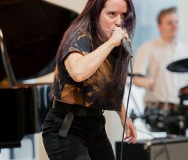 Female singer with purple-dyed hair singing into a microphone on stage, with a grand piano in the background.