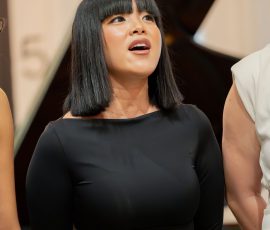 Female singer with black hair in a black dress, singing on stage near a grand piano.