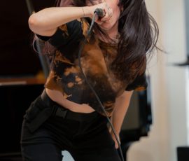 Female singer performing into a microphone, hair flying, wearing a tie-dye top and black pants on a stage-like setting.