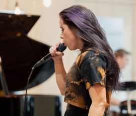 Female singer mid-performance, holding a microphone beside a grand piano in a softly lit room.