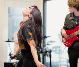 Female vocalist singing into a handheld microphone, wearing black clothing, with a red-bass guitarist in the background during a performance.