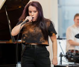 Female singer with purple-streaked hair singing into a microphone in a rehearsal space, a grand piano behind her.