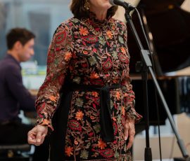 Woman singer wearing a colorful floral dress and red glasses, singing into a microphone with a pianist in the background.