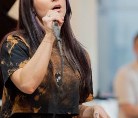 Female singer with long dark hair performing into a microphone on stage under warm lighting.