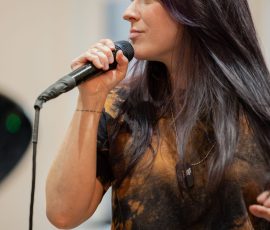 Woman singing into a handheld microphone, photographed in profile indoors, wearing a tie-dye shirt with long dark hair.