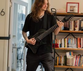 Person with long hair plays a black electric guitar in a home studio, surrounded by a bookshelf and gear behind them.