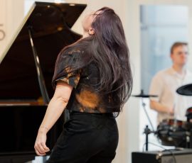 Woman with long dark hair leaning back while playing a grand piano in a bright room, with a drummer in the background.