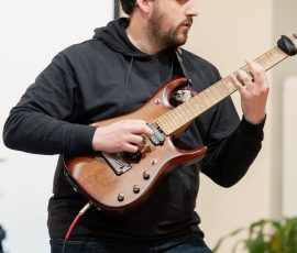Male guitarist wearing a black hoodie playing a brown electric guitar on stage or indoors, focused on fretboard artfully.