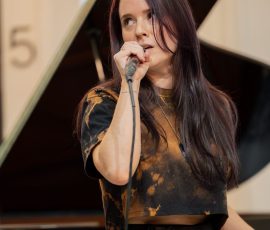 Female singer with long dark hair singing into a microphone on a stage, a grand piano visible in the background.