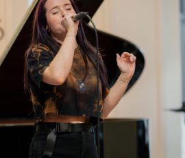 Female singer with purple-dyed hair singing into a microphone, eyes closed, with a grand piano in the background.