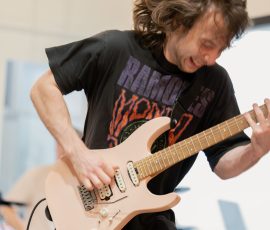 Male guitarist in a black T-shirt and brown pants plays a pale pink electric guitar on stage, hair swinging with the motion of the performance.