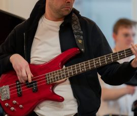 Man in a black cap and jacket playing a red bass guitar, focused on the fretboard during a rehearsal.